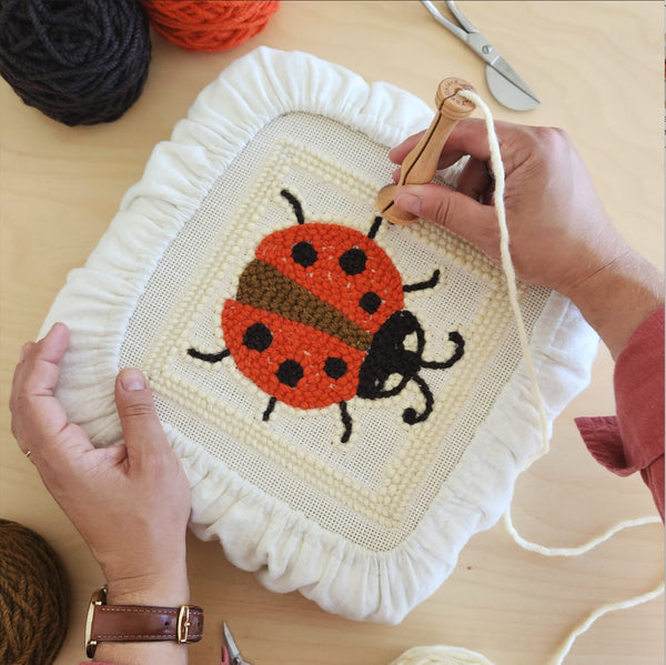 above the table view of two hands punching a 6" ladybug pattern with wool yarn