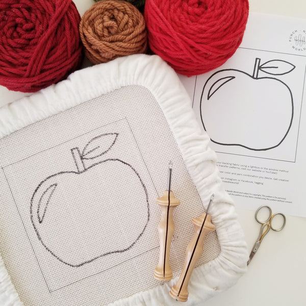 An apple design traced onto monkscloth and stretched over a frame, prepared for punching. Oxford Punch Needles, yarn and small scissors laying nearby.